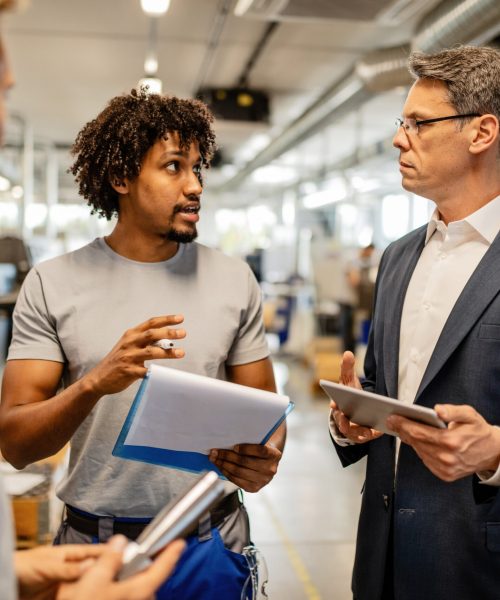 Mid adult engineer using touchpad and communicating with African American worker about product reports in industrial building.